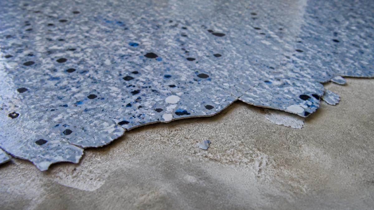 Close-up of concrete garage floor showing peeling epoxy coating with visible separation, flaking edges, and exposed concrete substrate in natural daylight